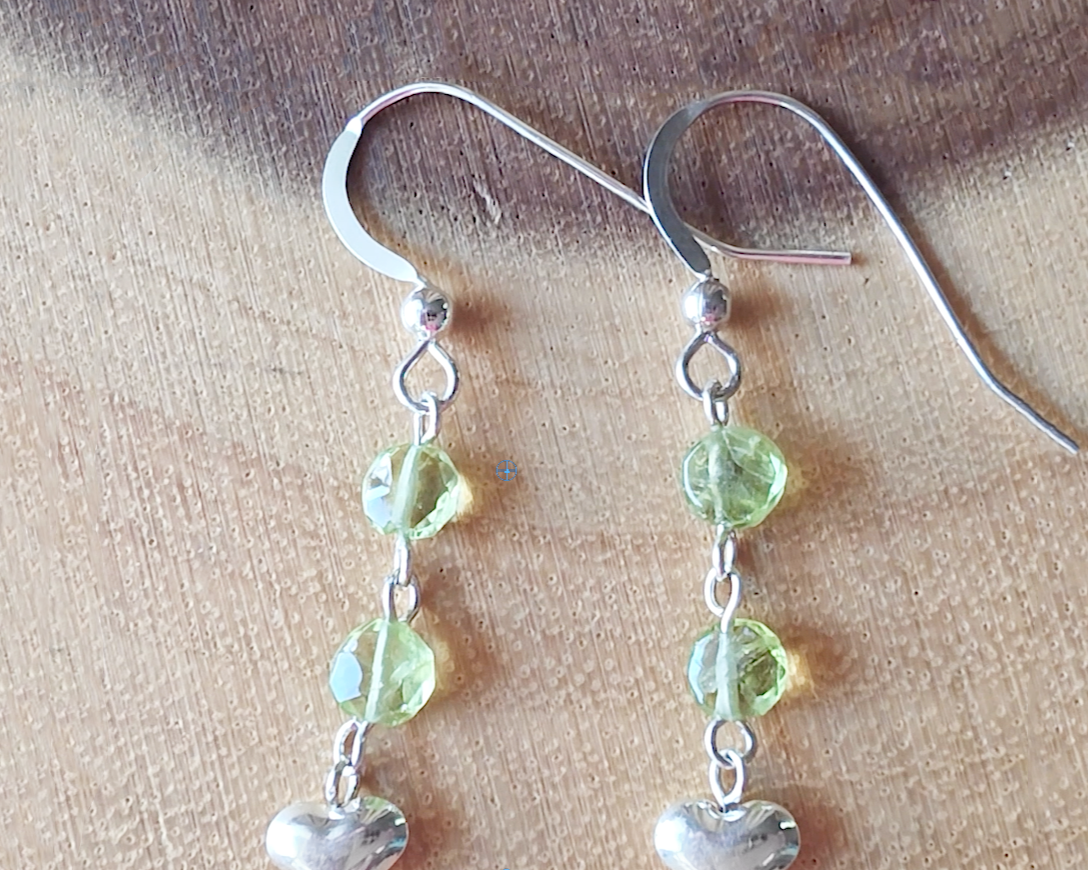 A pair of long sparkly Peridot Sterling silver earrings with two round faceted green gemstones and silver hearts on the base dangling from french earring hooks on wood 