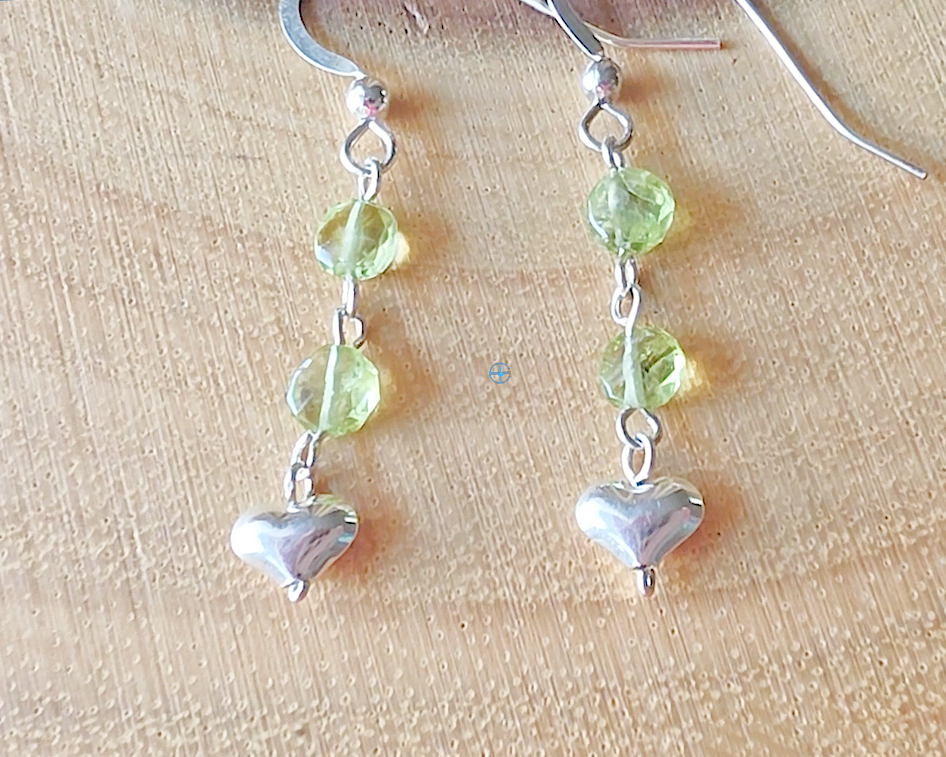 A pair of long sparkly Peridot Sterling silver earrings with two round faceted green gemstones and silver hearts on the base dangling from french earring hooks on wood 