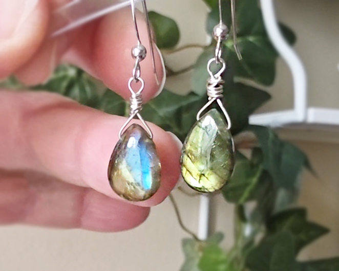 Handmade Labradorite drop shaped earrings with colorful flashes of blue and gold on sterling silver french hooks with plants in the background on hand