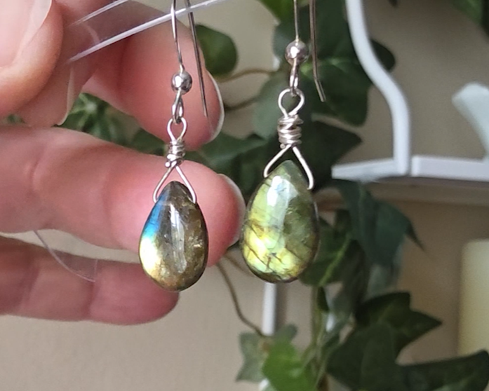 Handmade Labradorite drop shaped earrings with colorful flashes of blue and gold on sterling silver french hooks with plants in the background on hand