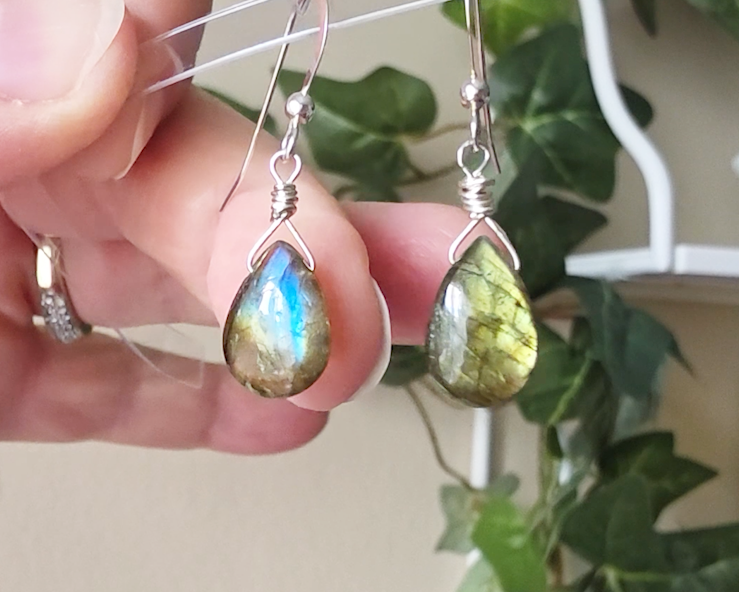 Handmade Labradorite drop shaped earrings with colorful flashes of blue and gold on sterling silver french hooks with plants in the background on hand