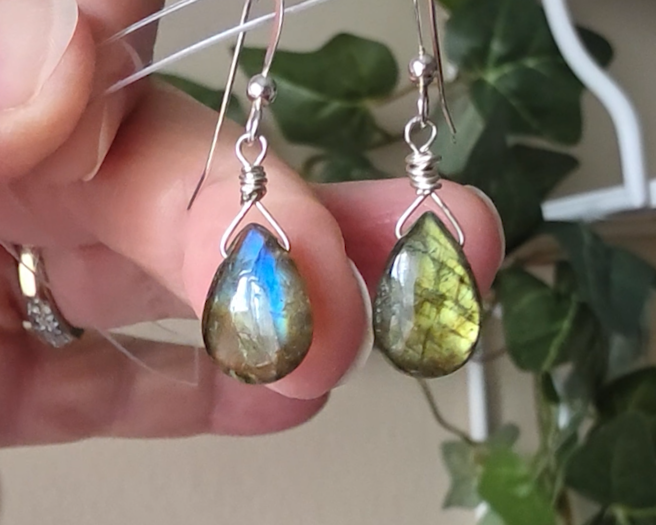 Handmade Labradorite drop shaped earrings with colorful flashes of blue and gold on sterling silver french hooks with plants in the background on hand