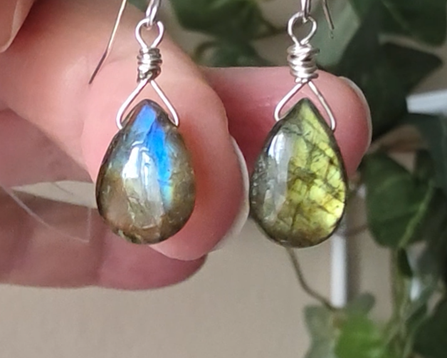 Handmade Labradorite drop shaped earrings with colorful flashes of blue and gold on sterling silver french hooks with plants in the background on hand