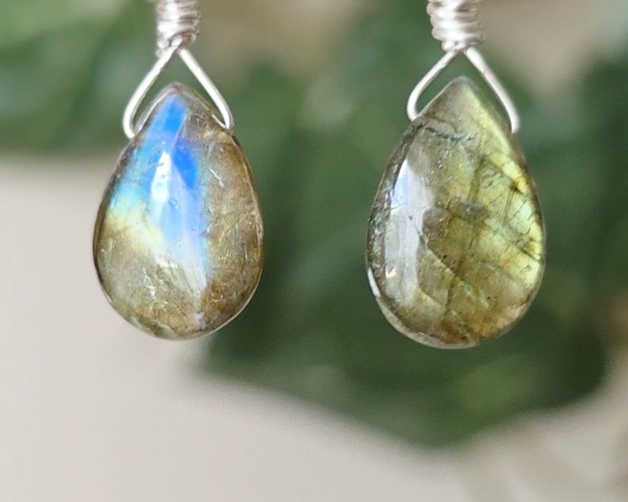 Handmade Labradorite drop shaped earrings with colorful flashes of blue and gold on sterling silver french hooks with plants in the background on hand