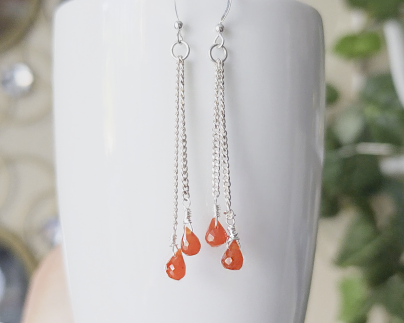 A pair of handmade long dangle earrings with orange faceted Carnelian drop shaped stones dangling from Sterling Silver chain on white background