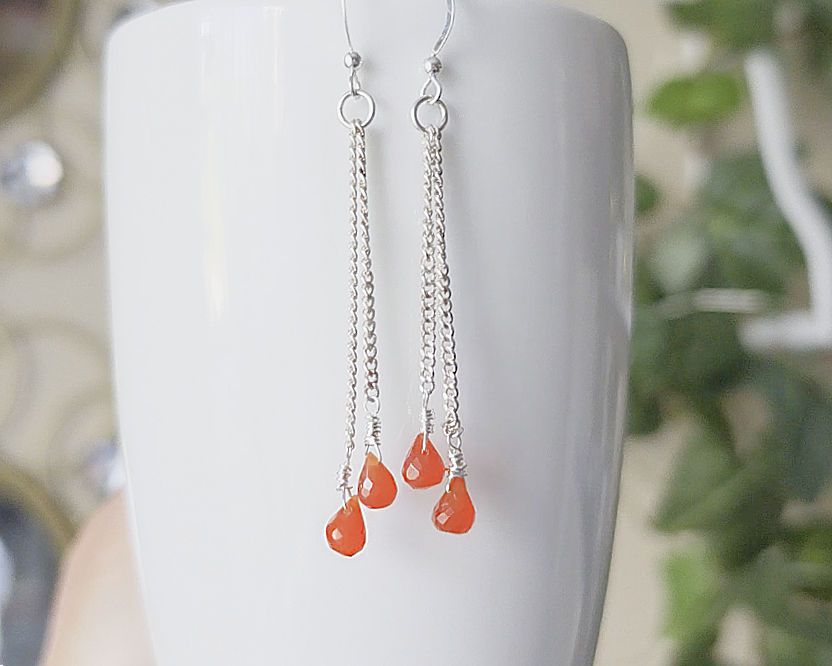 A pair of handmade long dangle earrings with orange faceted Carnelian drop shaped stones dangling from Sterling Silver chain on white background