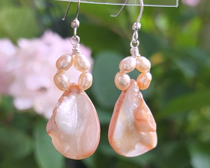 A pair of Pale Pink Freshwater Cultured Peral and Vintage Shell Drop Earrings on Sterling Silver French earring hooks dangling in front of hand with flowers in background.