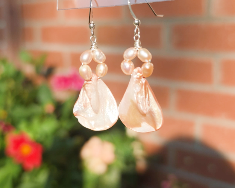 A pair of Pale Pink Freshwater Cultured Peral and Vintage Shell Drop Earrings on Sterling Silver French earring hooks dangling in front of hand with garden in background.