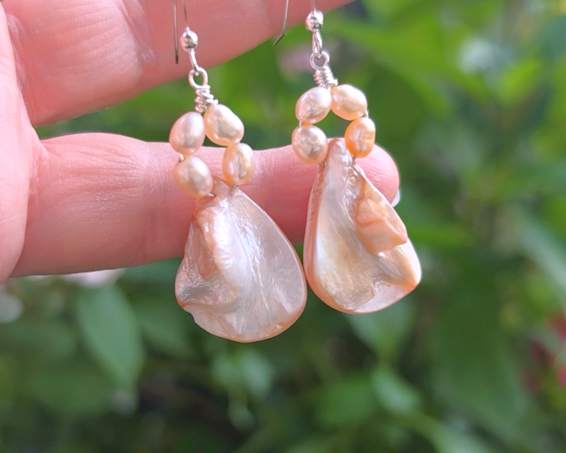A pair of Pale Pink Freshwater Cultured Peral and Vintage Shell Drop Earrings on Sterling Silver French earring hooks dangling in front of hand with garden in background.