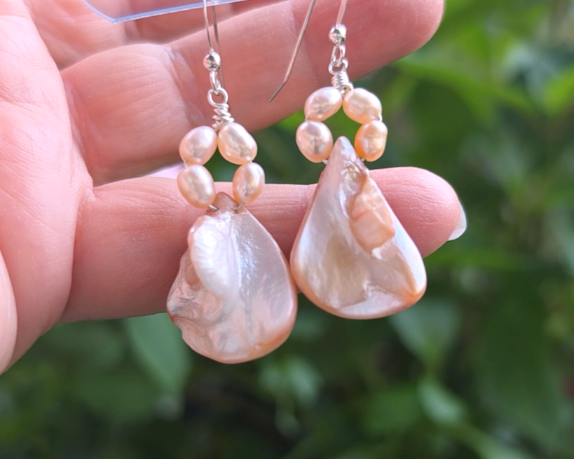 A pair of Pale Pink Freshwater Cultured Peral and Vintage Shell Drop Earrings on Sterling Silver French earring hooks dangling in front of hand with garden in background.