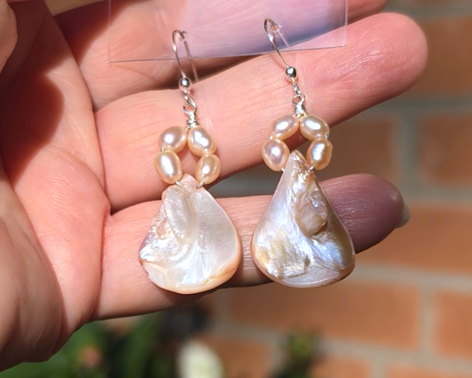 A pair of Pale Pink Freshwater Cultured Peral and Vintage Shell Drop Earrings on Sterling Silver French earring hooks dangling in front of hand with brick wall in background.