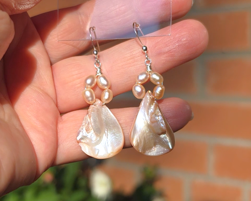 A pair of Pale Pink Freshwater Cultured Peral and Vintage Shell Drop Earrings on Sterling Silver French earring hooks dangling in front of hand with brick wall in background.
