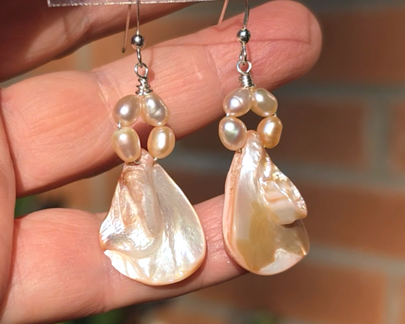 A pair of Pale Pink Freshwater Cultured Peral and Vintage Shell Drop Earrings on Sterling Silver French earring hooks dangling in front of hand with brick wall in background.