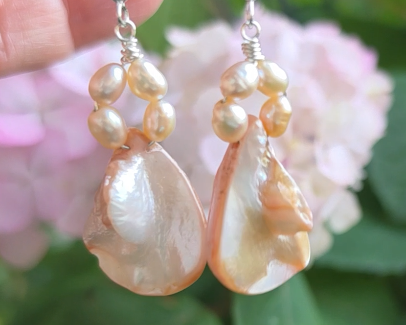 A pair of Pale Pink Freshwater Cultured Peral and Vintage Shell Drop Earrings on Sterling Silver French earring hooks dangling in front of hand with garden in background.