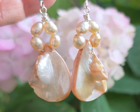 A pair of Pale Pink Freshwater Cultured Peral and Vintage Shell Drop Earrings on Sterling Silver French earring hooks dangling in front of hand with garden in background.