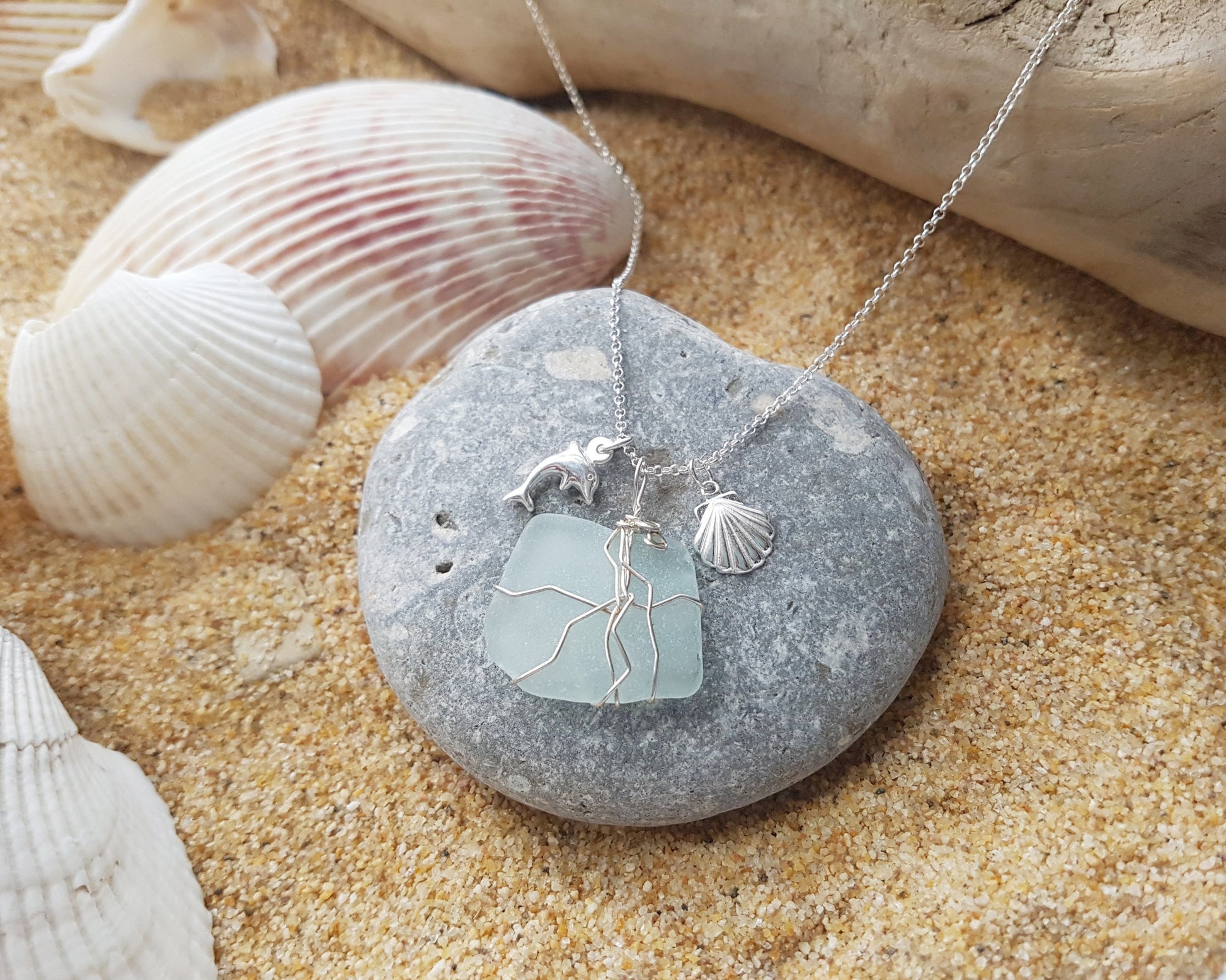 Handmade pale Blue Sea Glass pendant wire wrapped with Sterling silver wire with dolphin pendant and shell pendant on chain on grey rock and sand, shells and wood