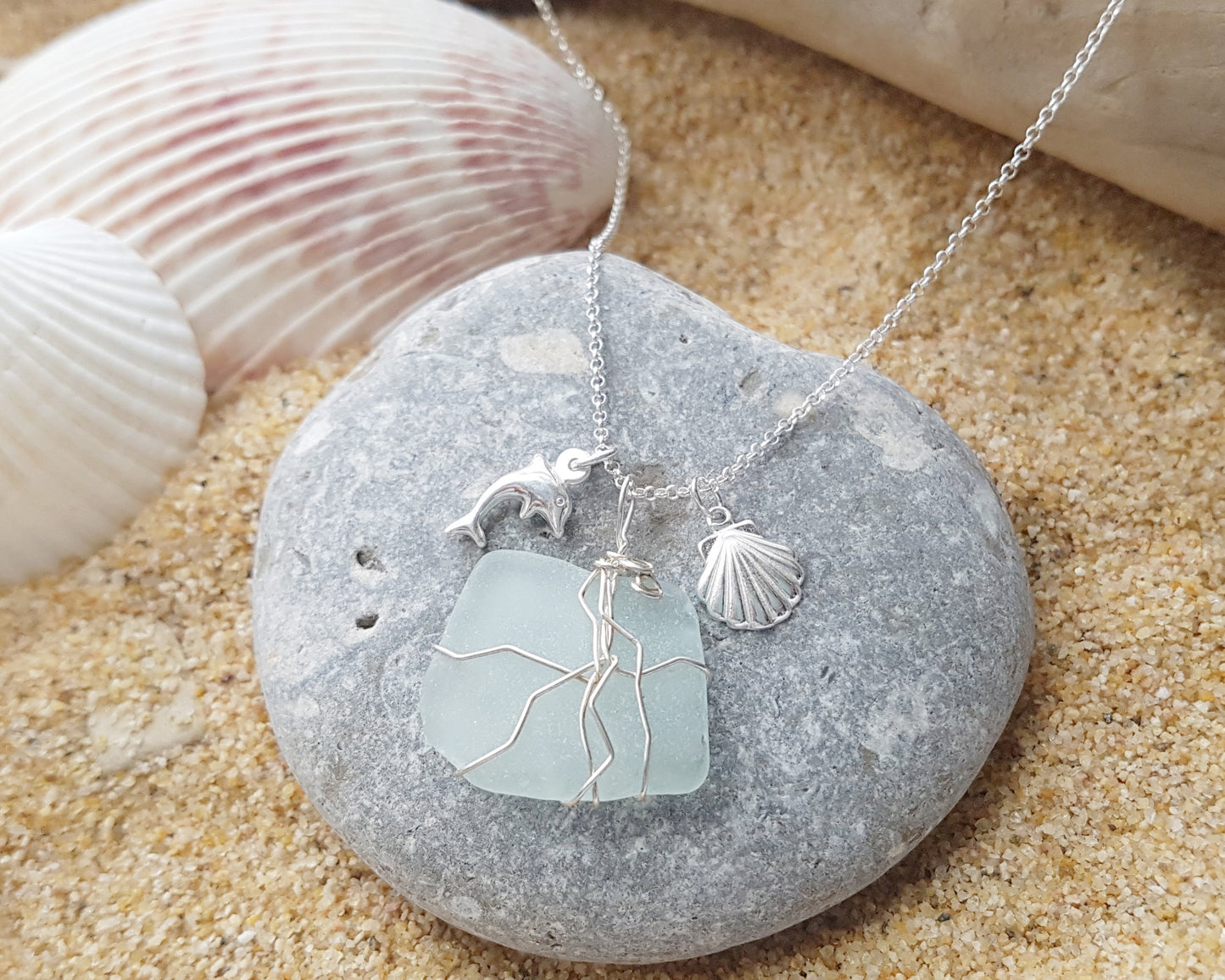 Handmade pale Blue Sea Glass pendant wire wrapped with Sterling silver wire with dolphin pendant and shell pendant on chain on grey rock and sand, shells and wood