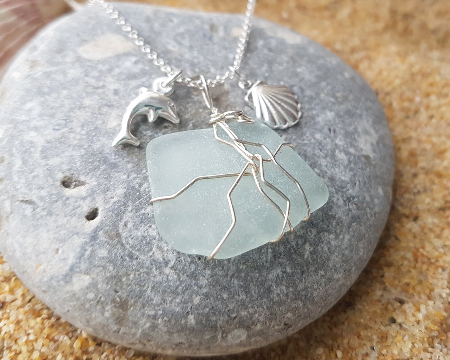 Handmade pale Blue Sea Glass pendant wire wrapped with Sterling silver wire with dolphin pendant and shell pendant on chain on grey rock and sand 