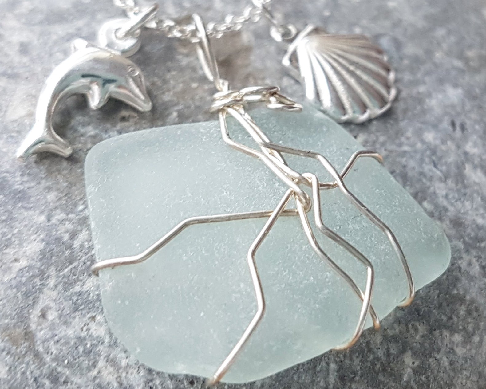 Handmade pale Blue Sea Glass pendant wire wrapped with Sterling silver wire with dolphin pendant and shell pendant on chain on grey rock 