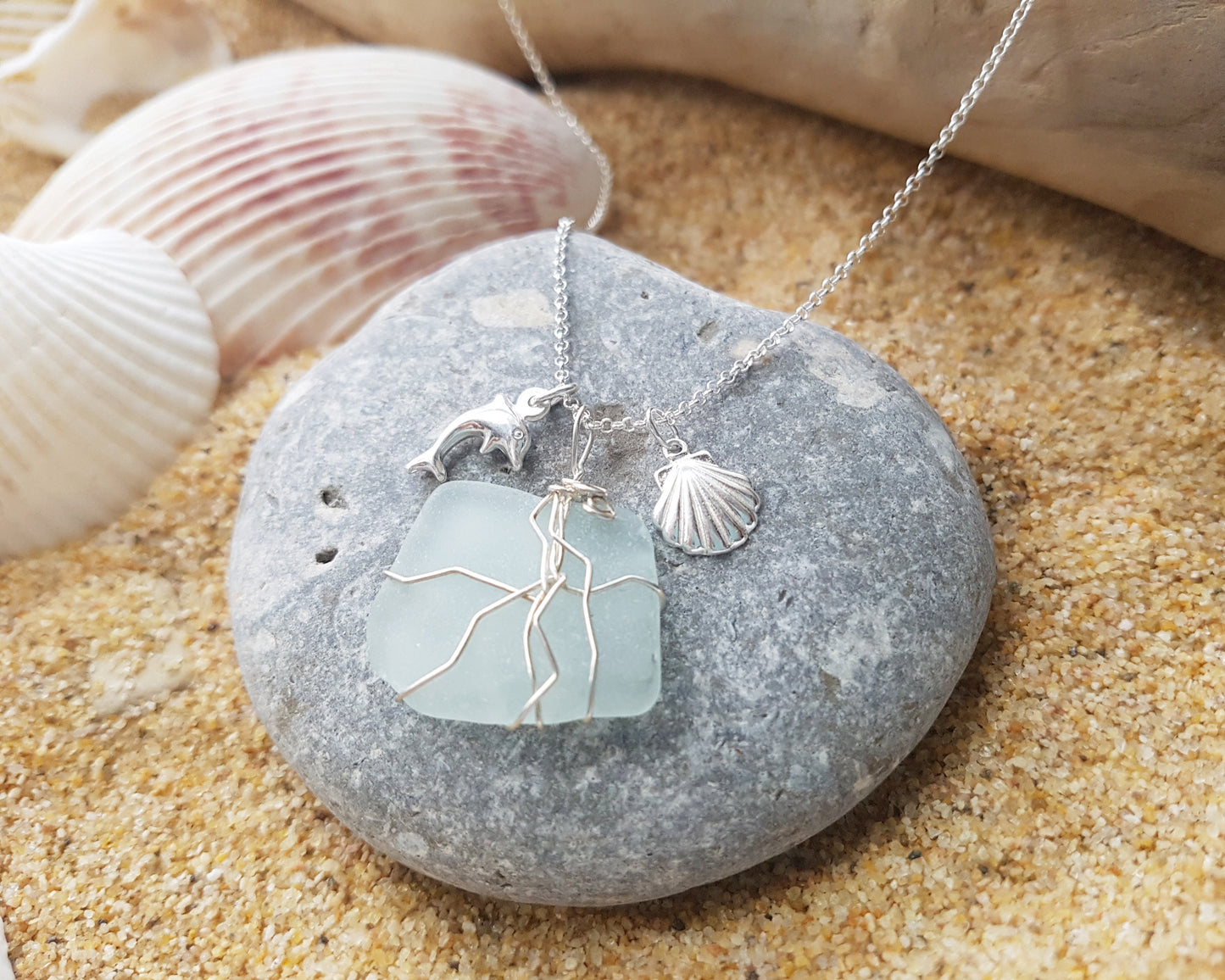 Handmade pale Blue Sea Glass pendant wire wrapped with Sterling silver wire with dolphin pendant and shell pendant on chain on grey rock and sand 