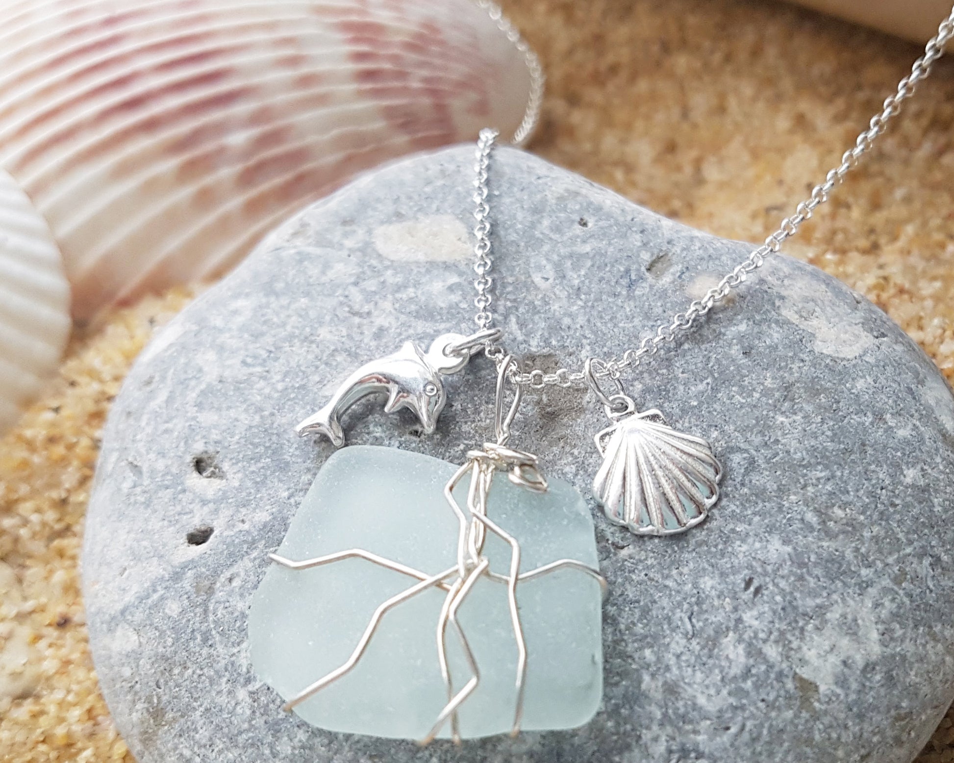 Handmade pale Blue Sea Glass pendant wire wrapped with Sterling silver wire with dolphin pendant and shell pendant on chain on grey rock and sand 