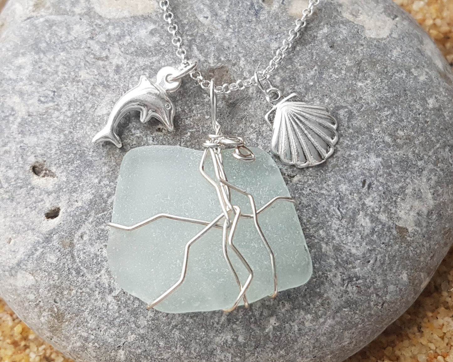 Handmade pale Blue Sea Glass pendant wire wrapped with Sterling silver wire with dolphin pendant and shell pendant on chain on grey rock and sand 