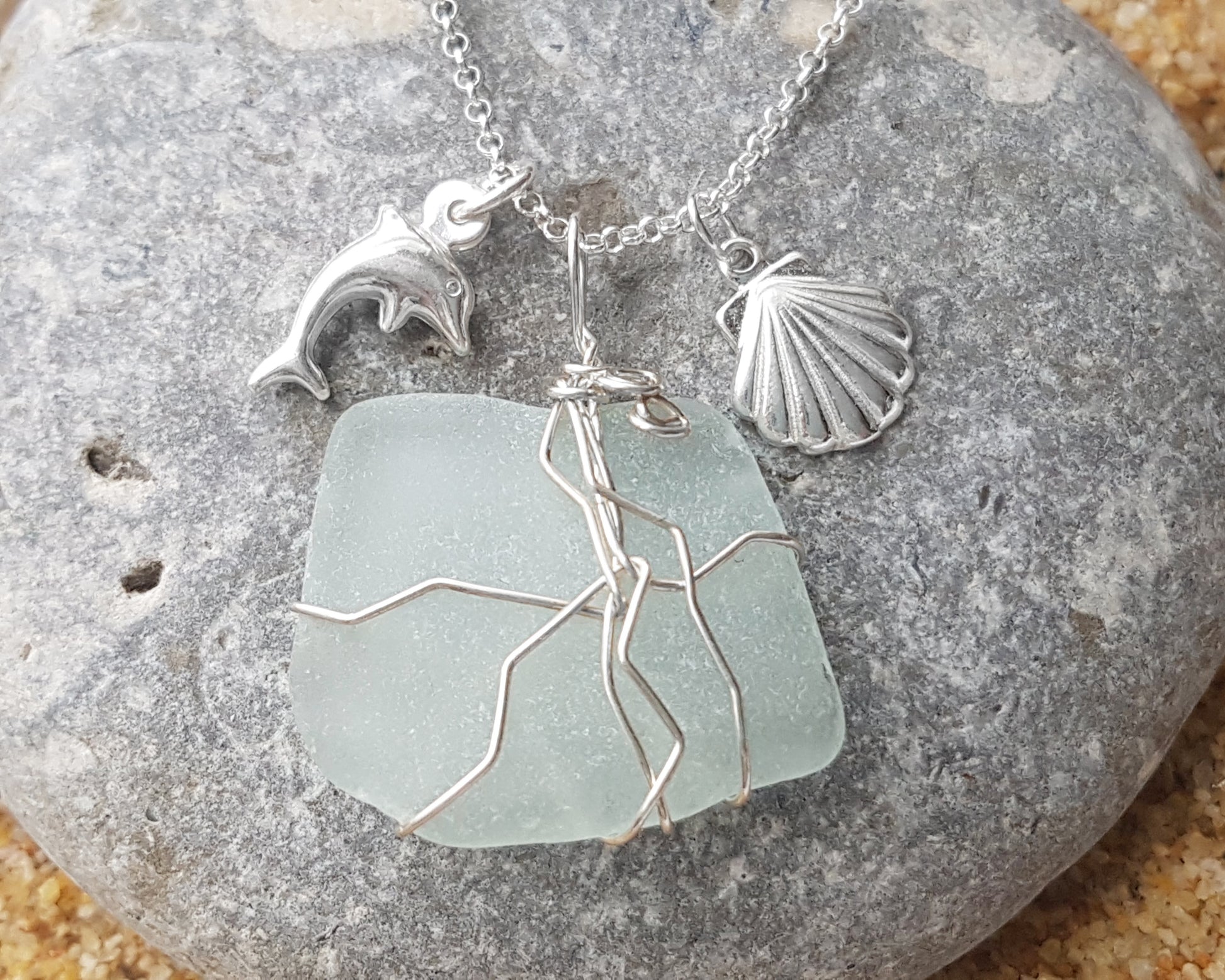 Handmade pale Blue Sea Glass pendant wire wrapped with Sterling silver wire with dolphin pendant and shell pendant on chain on grey rock and sand 