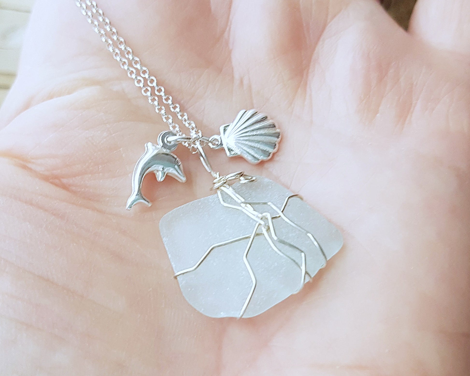 Handmade pale Blue Sea Glass pendant wire wrapped with Sterling silver wire with dolphin pendant and shell pendant on chain on grey rock and sand 