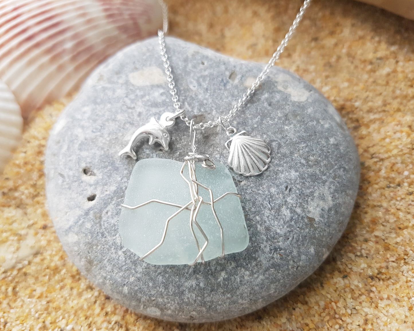 Handmade pale Blue Sea Glass pendant wire wrapped with Sterling silver wire with dolphin pendant and shell pendant on chain on grey rock and sand 