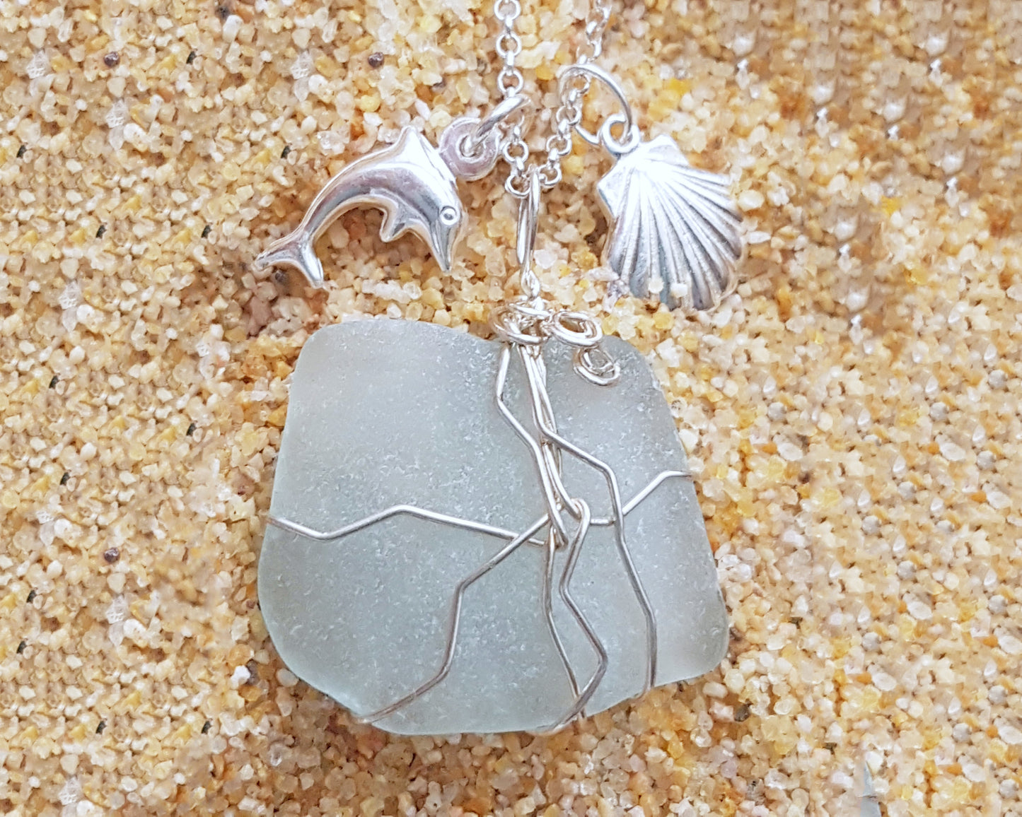 Handmade pale Blue Sea Glass pendant wire wrapped with Sterling silver wire with dolphin pendant and shell pendant on chain on sand 