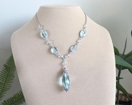 Art Deco style Aquamarine necklace made with large faceted, sparkly pale aqua blue translucent synthetic Aquamarine stones and Sterling Silver beads and chain on beige display and plants in background 