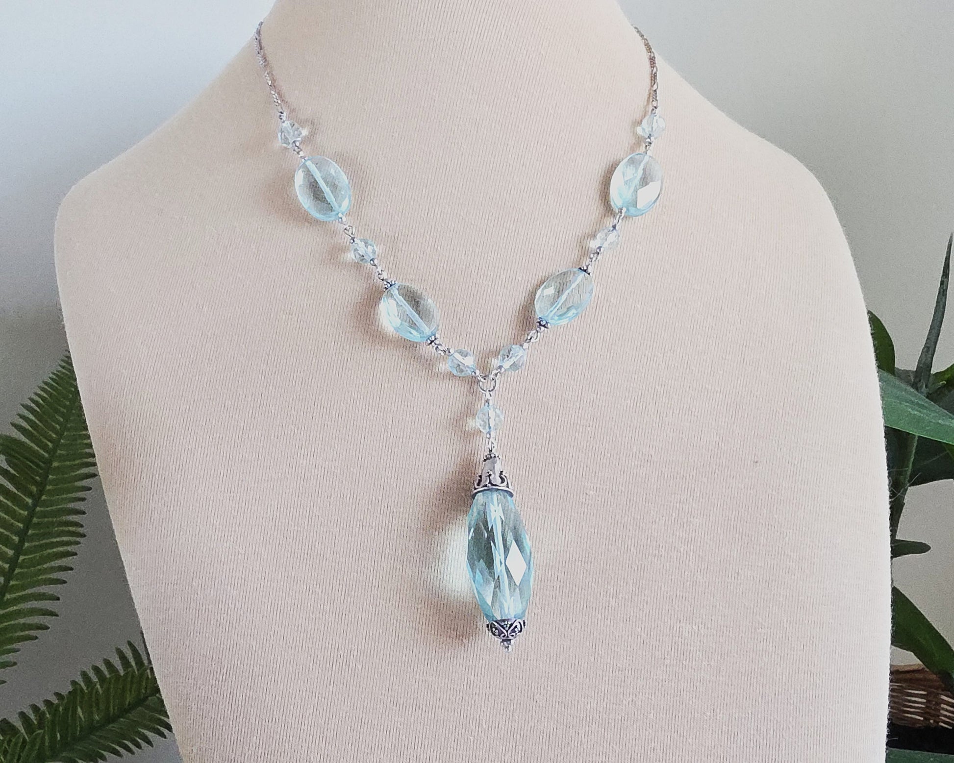 Art Deco style Aquamarine necklace made with large faceted, sparkly pale aqua blue translucent synthetic Aquamarine stones and Sterling Silver beads and chain on beige display and plants in background 