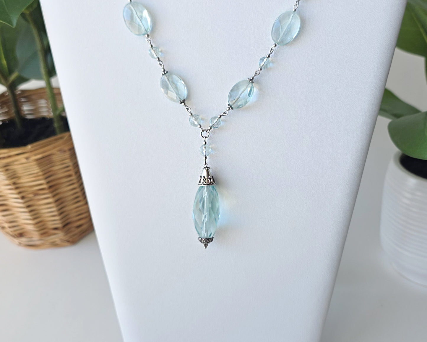 Art Deco style Aquamarine necklace made with large faceted, sparkly pale aqua blue translucent synthetic Aquamarine stones and Sterling Silver beads and chain on white background and plants 