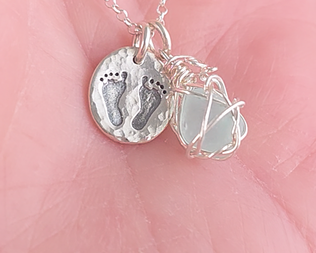 Pale Blue Sea Glass Pendant with round Silver Pendant with Footprints on Chain displayed on hand.