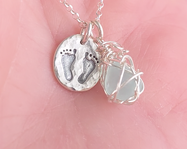 Pale Blue Sea Glass Pendant with round Silver Pendant with Footprints on Chain displayed on hand.