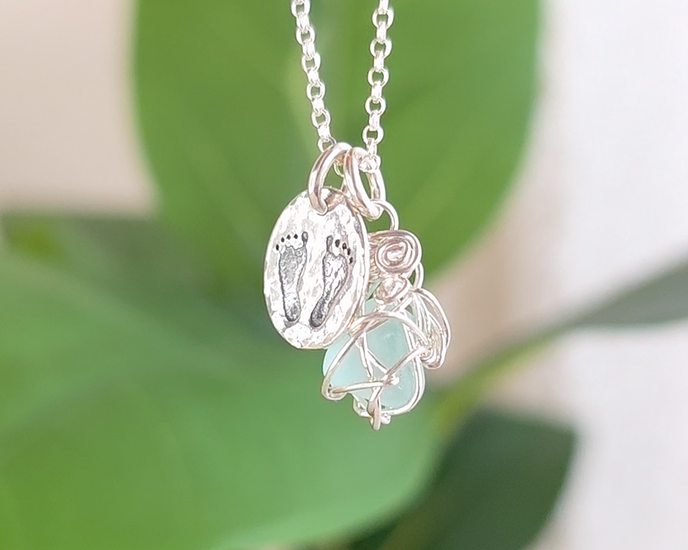 Pale Blue Sea Glass Pendant with round Silver Pendant with Footprints on Chain with green plants in the background 