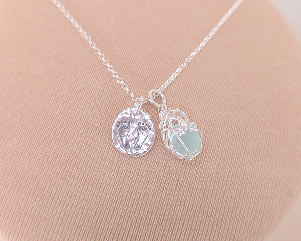 Pale Blue Sea Glass Pendant with round Silver Pendant with Footprints on Chain displayed on beige background