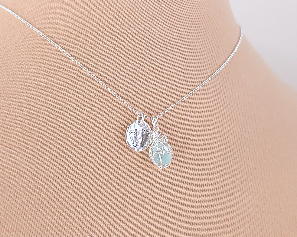 Pale Blue Sea Glass Pendant with round Silver Pendant with Footprints on Chain displayed on beige background 