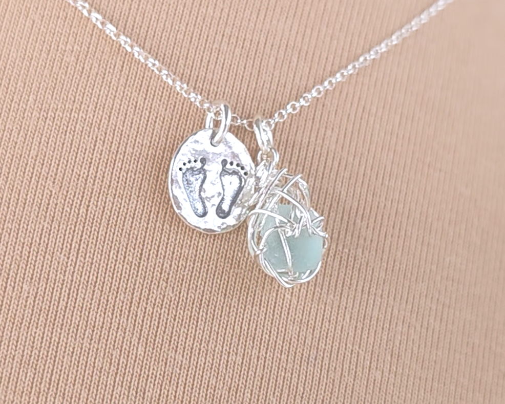Pale Blue Sea Glass Pendant with round Silver Pendant with Footprints on Chain displayed on beige background