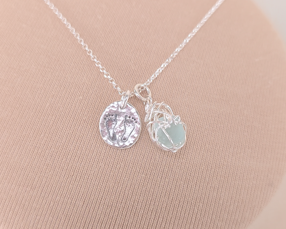 Pale Blue Sea Glass Pendant with round Silver Pendant with Footprints on Chain displayed on beige background