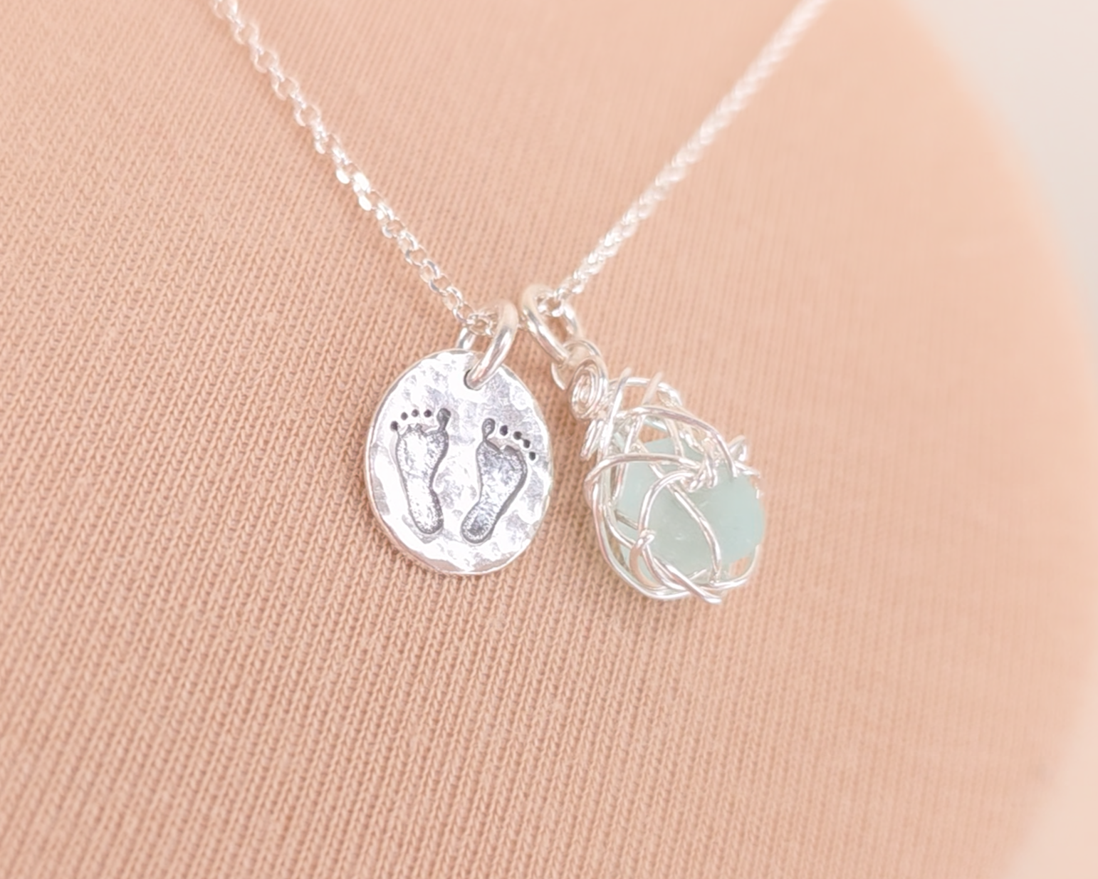 Pale Blue Sea Glass Pendant with round Silver Pendant with Footprints on Chain displayed on beige background 