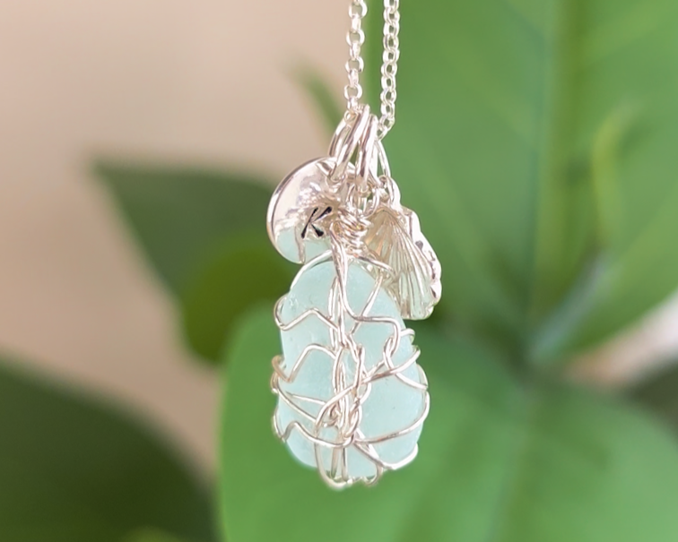 Silver Sea Glass Necklace with Aqua blue Sea Glass wire wrapped with shell & initial pendants on chain with green plants in the background 