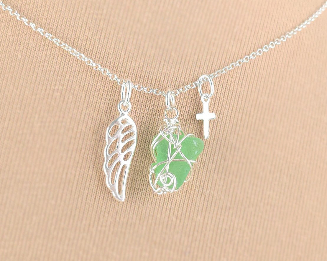 Silver necklace with green sea glass wire wrapped pendant with a silver wing and small cross on chain with beige background