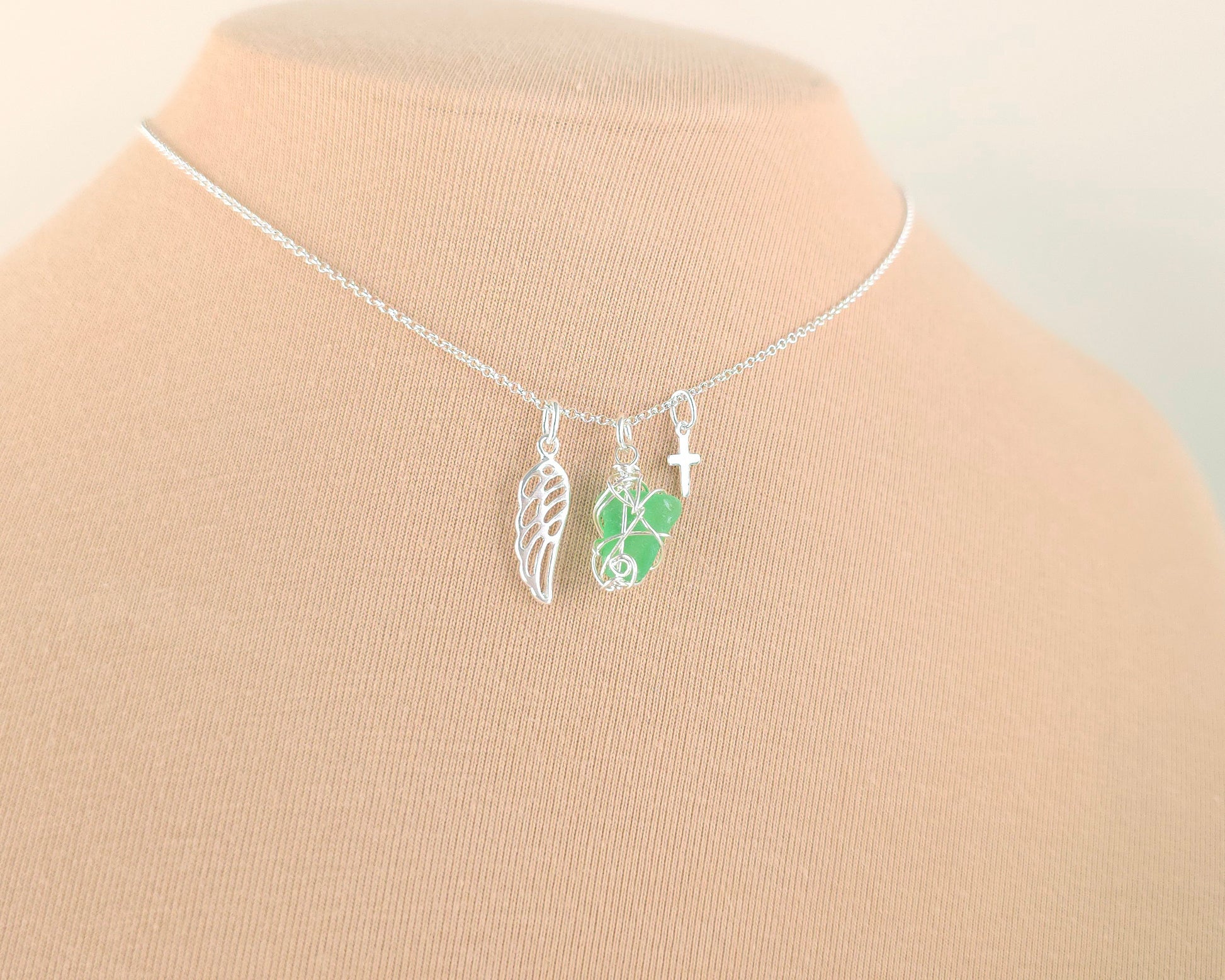 Silver necklace with green sea glass wire wrapped pendant with a silver angel wing and small cross on chain with beige background