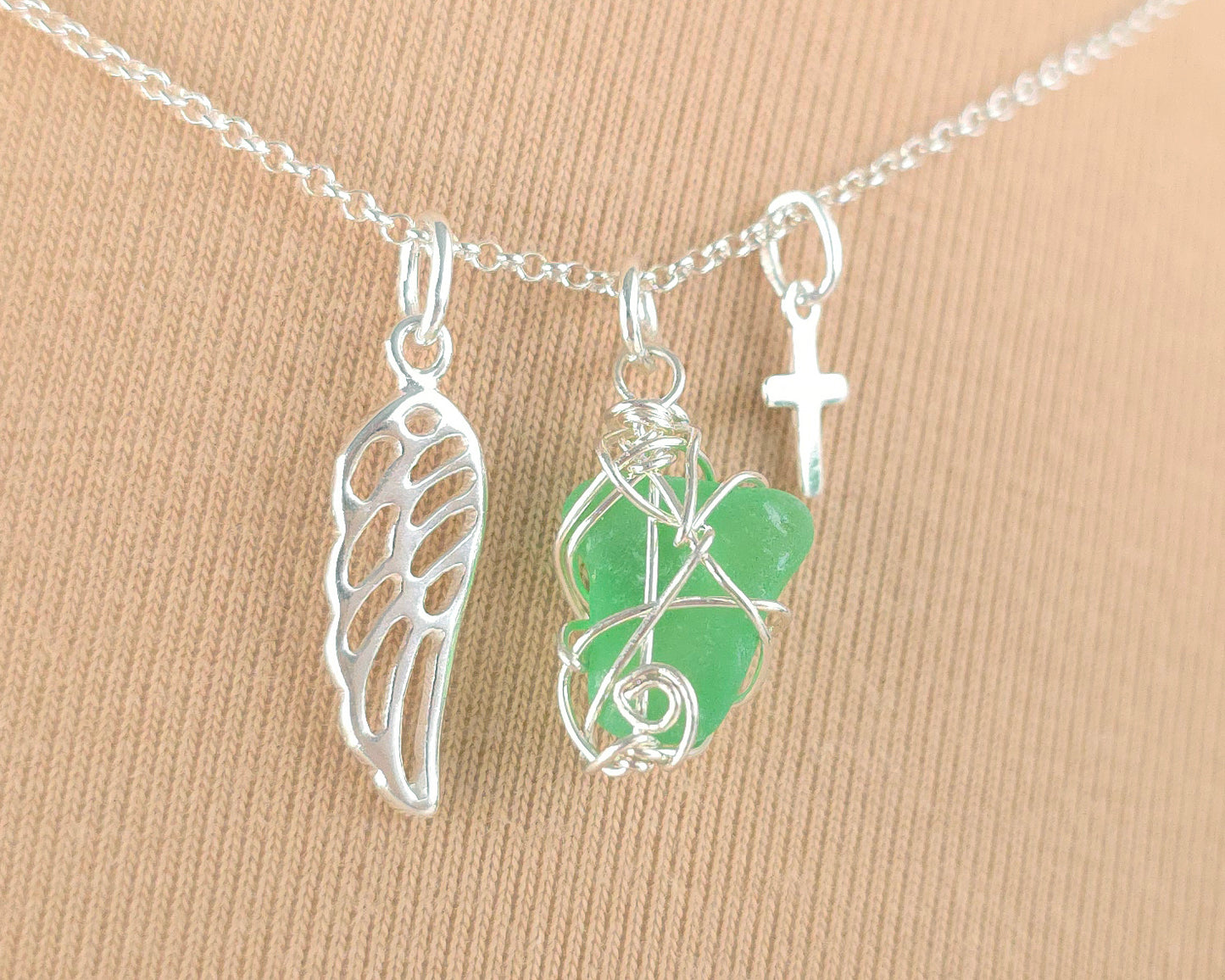 Silver necklace with green sea glass wire wrapped pendant with a silver angel wing and small cross on chain with beige background