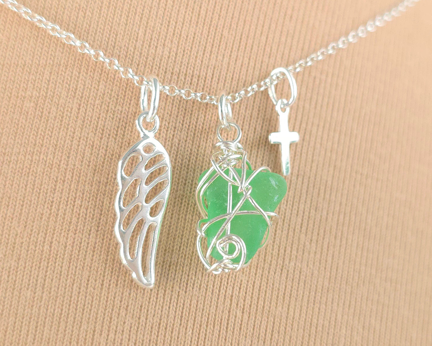 Silver necklace with green sea glass wire wrapped pendant with a silver angel wing and small cross on chain with beige background