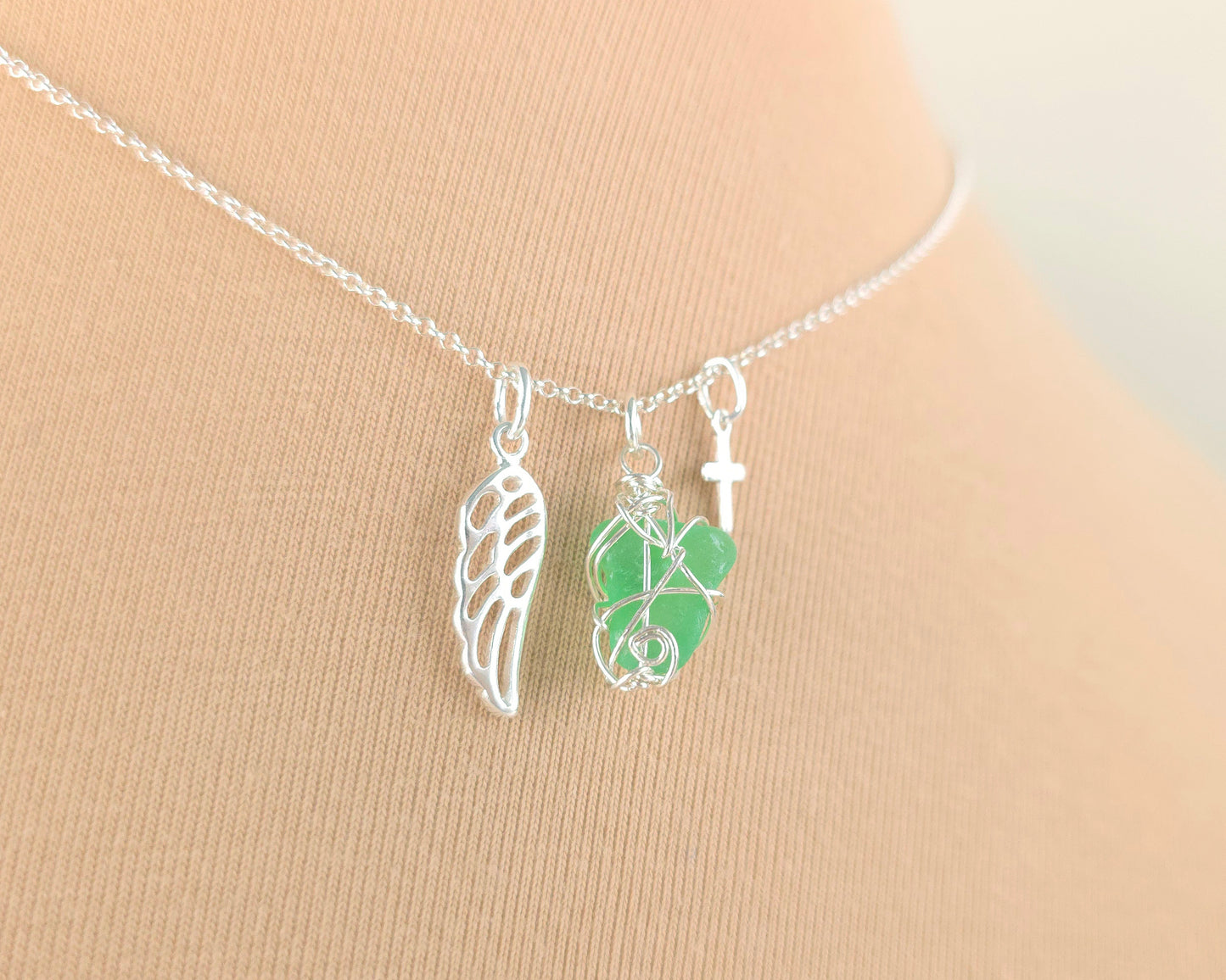 Silver necklace with green sea glass wire wrapped pendant with a silver angel wing and small cross on chain with beige background