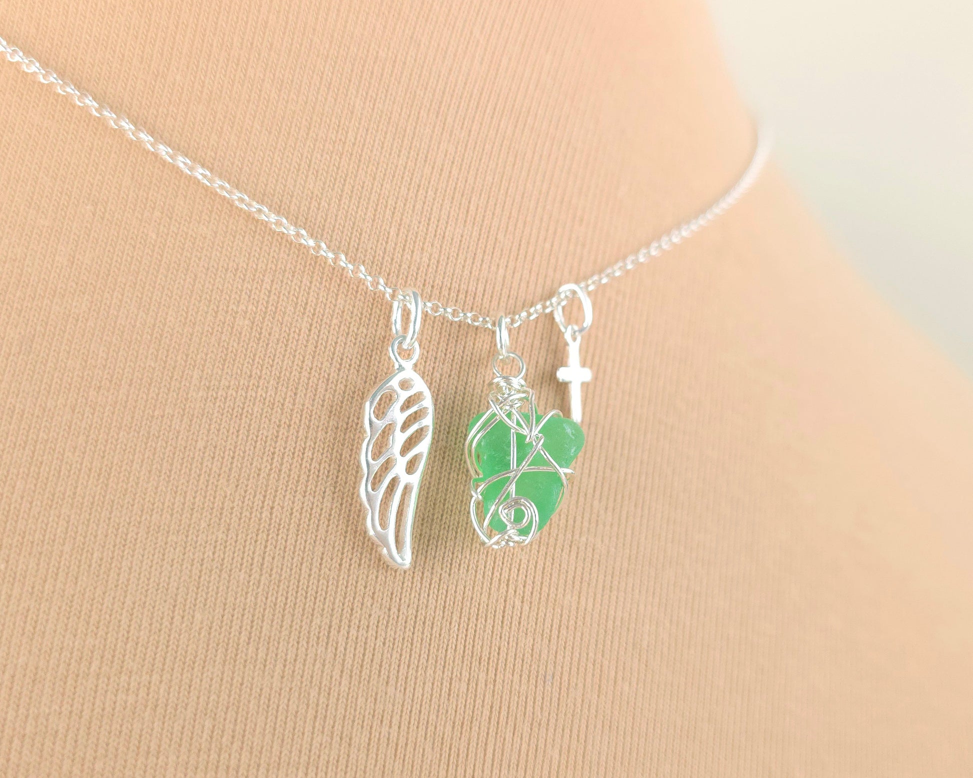 Silver necklace with green sea glass wire wrapped pendant with a silver angel wing and small cross on chain with beige background