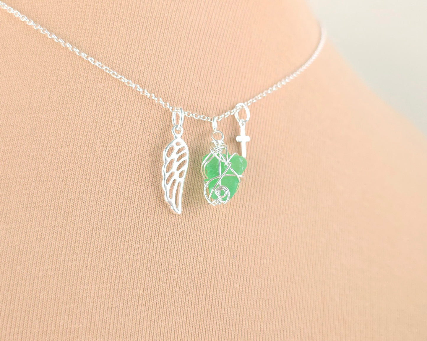 Silver necklace with green sea glass wire wrapped pendant with a silver angel wing and small cross on chain with beige background