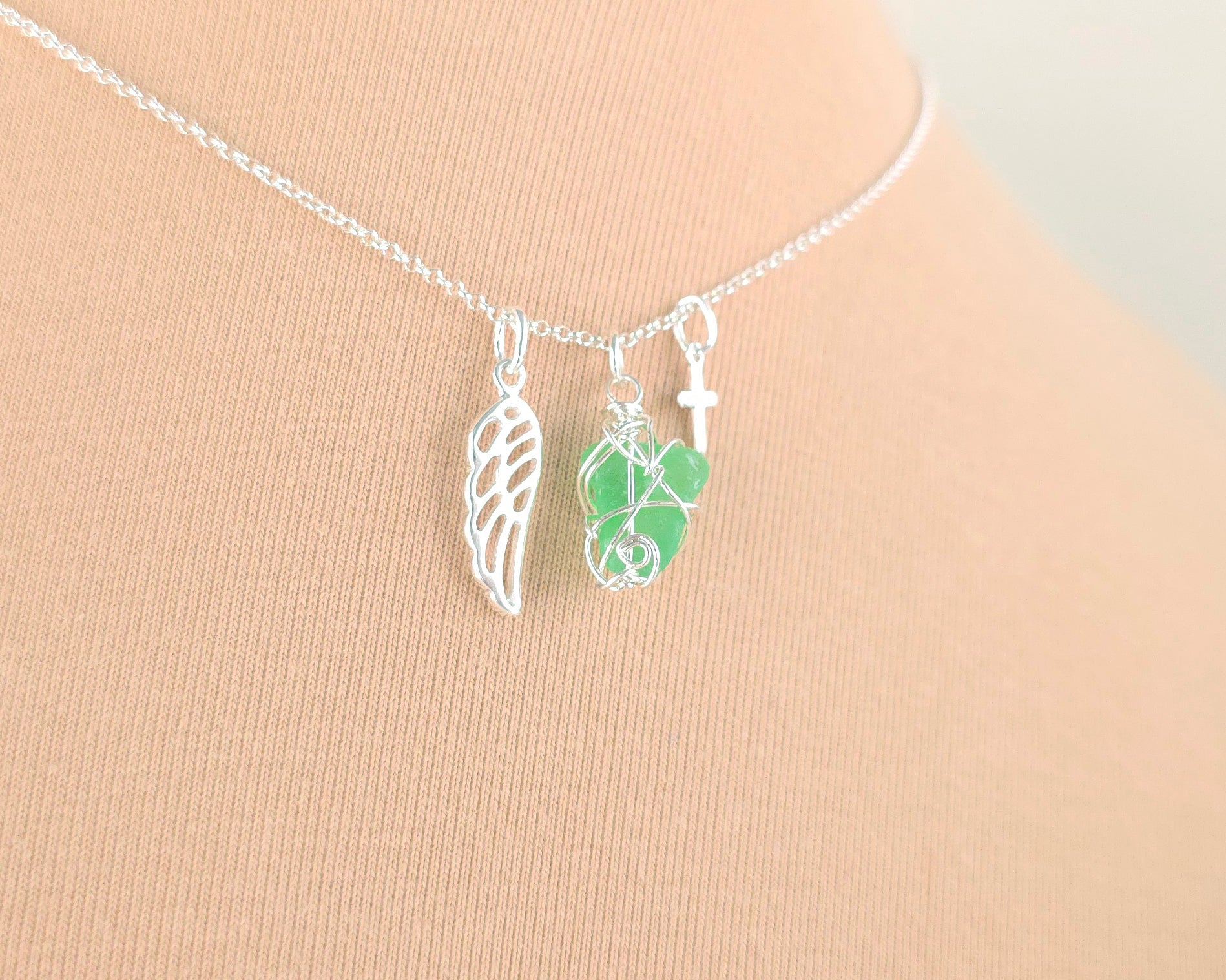Silver necklace with green sea glass wire wrapped pendant with a silver angel wing and small cross on chain with beige background